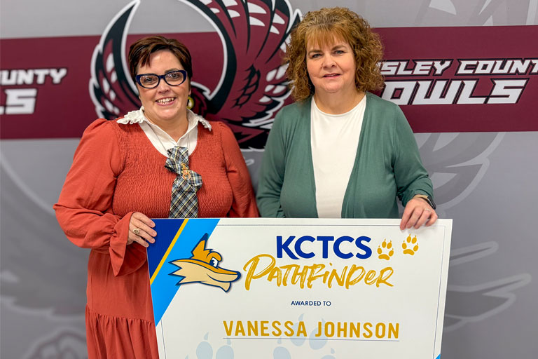 Two women standing side by side, holding a large award certificate. The certificate reads "KCTCS Pathfinder" with a fox logo and paw prints, and is awarded to "Vanessa Johnson." The background features a maroon and gray banner with the text "Wesley County Owls" and a stylized owl logo.